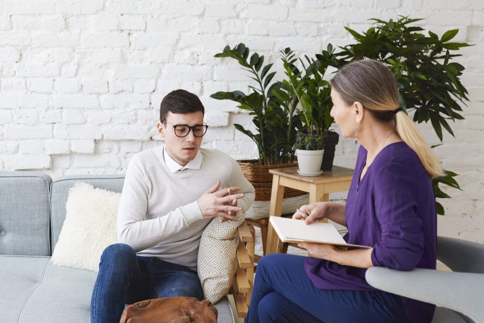 candid-shot-young-man-glasses-talking-about-his-problems-psychological-therapy-session-sitting-coach-while-mature-female-psychologist-with-copybook-listening-him-making-notes-min